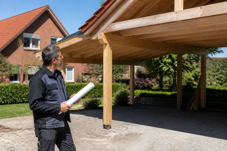Ein Mann prüft die Baupläne für ein neues Carport in einem deutschen Vorort an einem sonnigen Tag, während der Bau fortschreitet.
