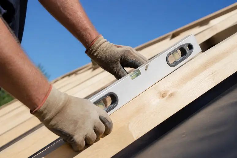Ein Handwerker prüft sorgfältig die Holzkonstruktion eines Daches, die als Grundlage für Bitumenschindeln dient, bei gutem Wetter.