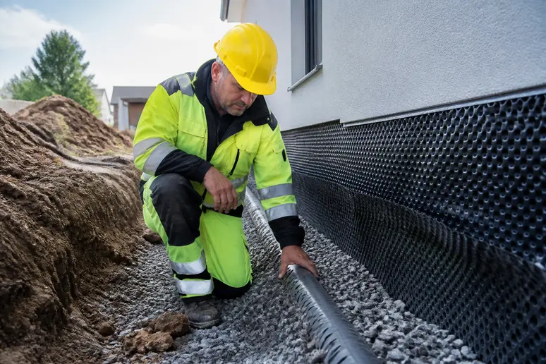 Ein Bauarbeiter überprüft ein Drainagerohr, das in Kies verlegt wurde, um ein Haus vor Feuchtigkeit zu schützen. Dieses Verfahren ist wichtig für die Gebäudesubstanz.