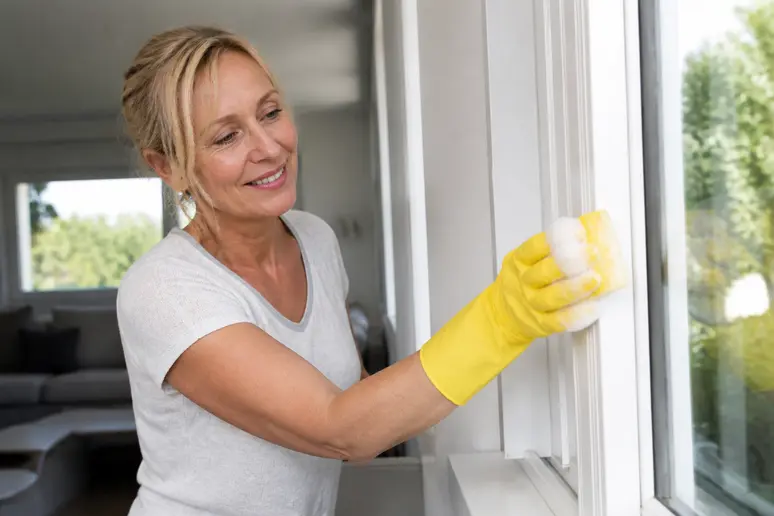 Eine Frau reinigt weiße Kunststofffensterrahmen mit einem Schwamm und Seifenwasser in einem hellen Wohnzimmer.