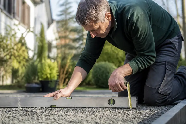 Ein Handwerker prüft mit Wasserwaage und Maßband den vorbereiteten Untergrund für Pflasterarbeiten im Garten.