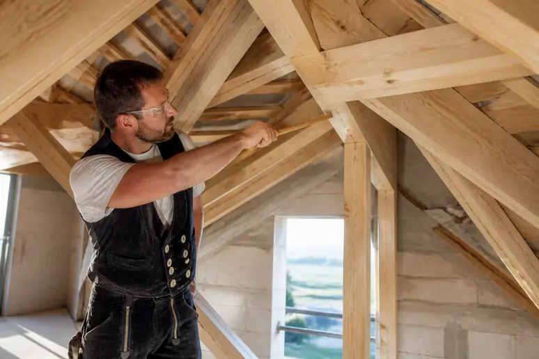Ein deutscher Zimmermann inspiziert sorgfältig die Holzkonstruktion eines Sparrendachs in einem Neubau, achtet auf Details.