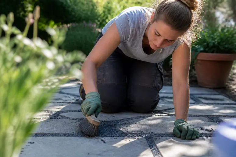 Frau kehrt Splitt in die Fugen von Terrassenplatten, um eine flexible und wasserdurchlässige Verbindung zu schaffen.