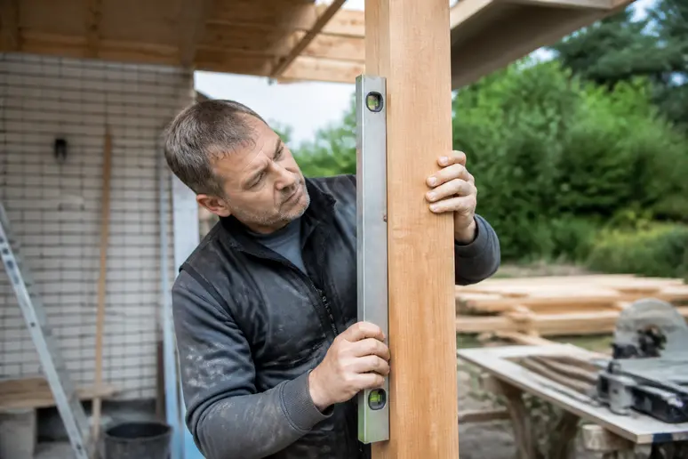 Ein Mann misst mit einer Wasserwaage die senkrechte Ausrichtung eines Holzpfostens für eine Vordachkonstruktion im Freien.