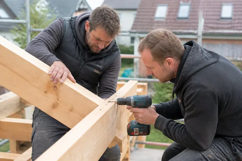 Zwei Männer montieren gemeinsam Holzbalken für ein selbstgebautes Vordach, ein dritter Mann im Hintergrund beobachtet.