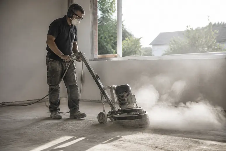 Handwerker mit Schleifmaschine trägt hohe Stellen auf Betonboden ab, Staubentwicklung sichtbar.