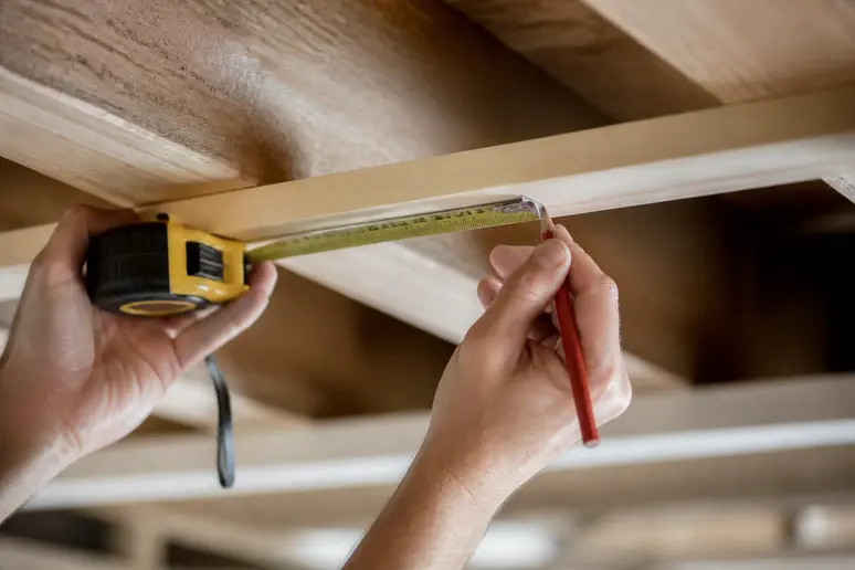 Hände markieren präzise Lattenabstände an Holzbalken für eine Deckenunterkonstruktion.