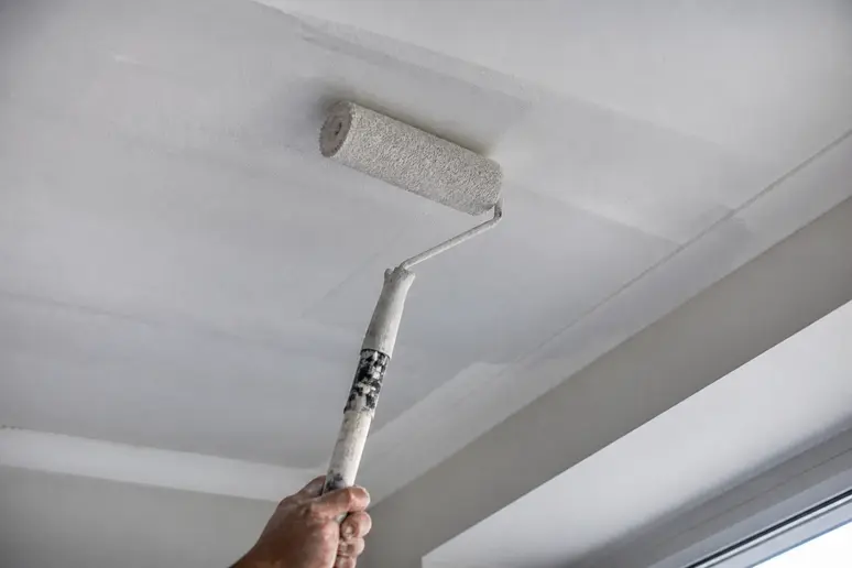 Hände streichen Decke quer zum Fensterlicht mit einer Farbrolle, überlappende Bahnen für ein gleichmäßiges Ergebnis.