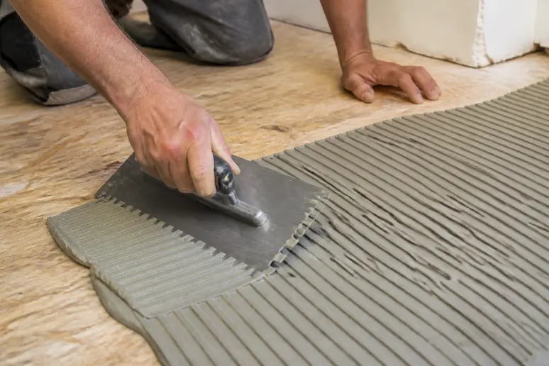 Fachmann trägt flexiblen Fliesenkleber mit Zahnspachtel auf Holzuntergrund auf, um Spannungen auszugleichen.
