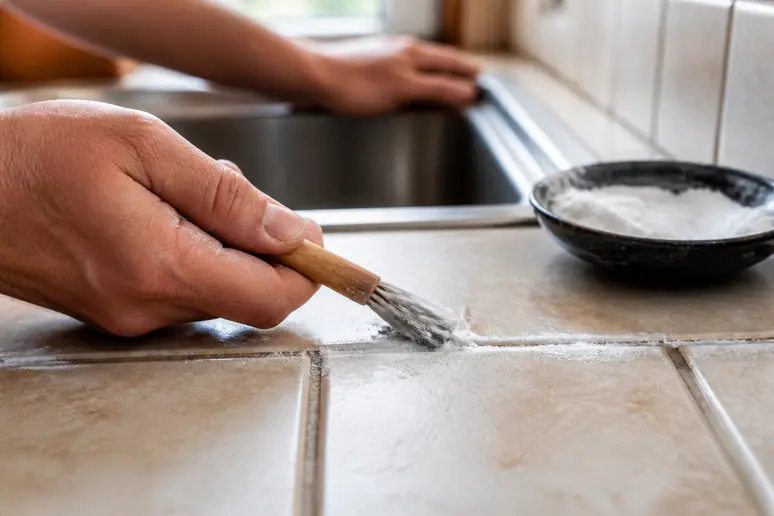 Hände tragen Natronpaste mit einer Bürste in Fliesenfugen im Küchenbereich auf.