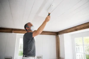 Holzdecke weiß streichen: Handwerker mit Roller bearbeitet sorgfältig die Decke im Innenraum für Renovierung.