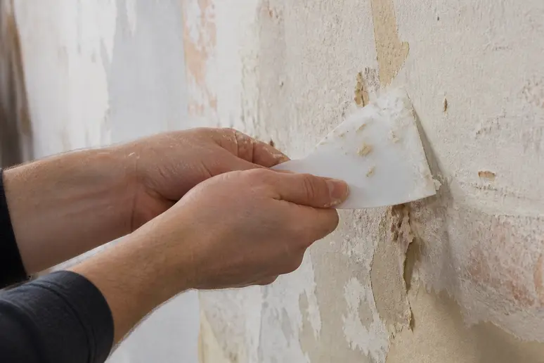 Hände mit Spachtel entfernen Kleisterreste von einer gestrichenen Wand.