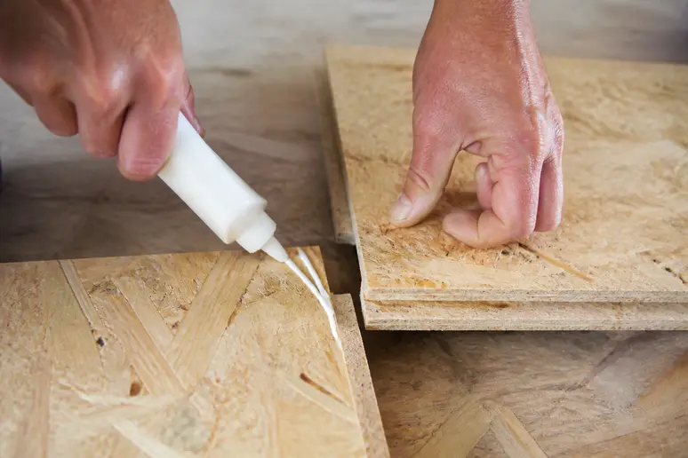 Hände tragen Holzleim in die Nut einer OSB-Platte auf, bevor die Feder einer anderen Platte eingesteckt wird.
