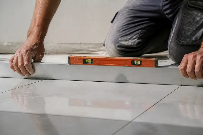Ein Handwerker prüft mit einer Richtlatte die Ebenheit eines Fliesenbodens auf sichtbare Unebenheiten.